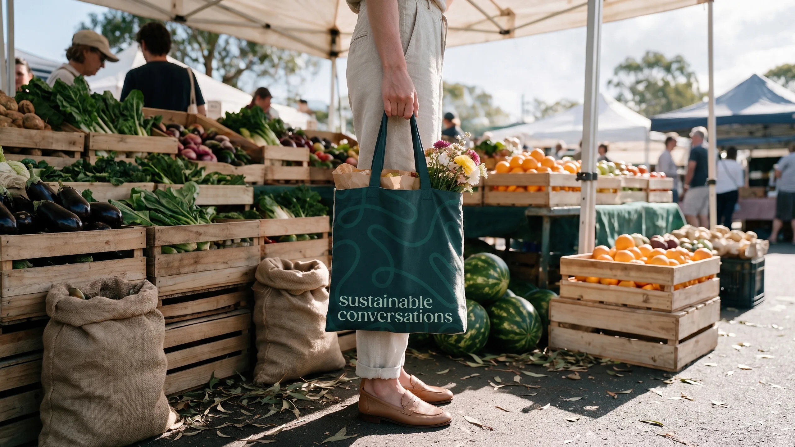 A person holds a dark teal tote bag with "sustainable conversations" printed on it at an outdoor market.
