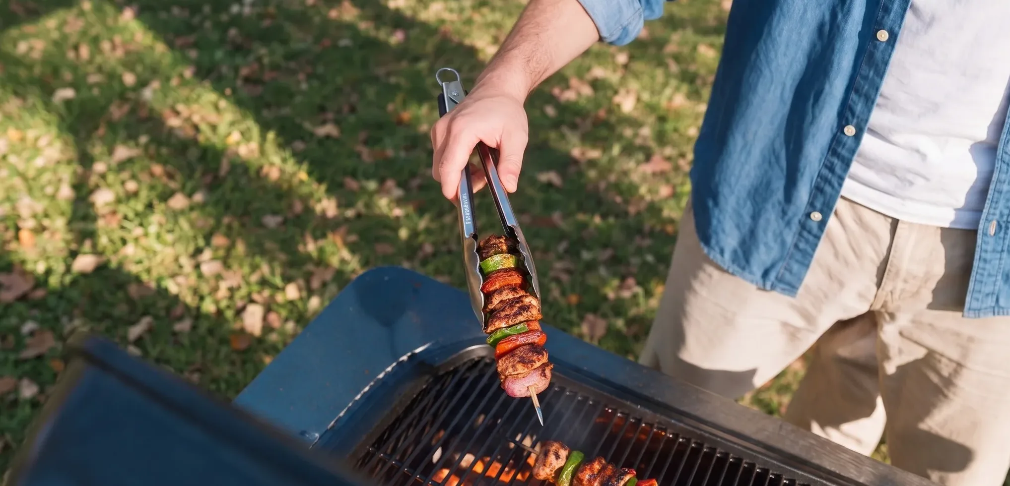 A person uses tongs to turn a skewer of meat and vegetables on a grill.