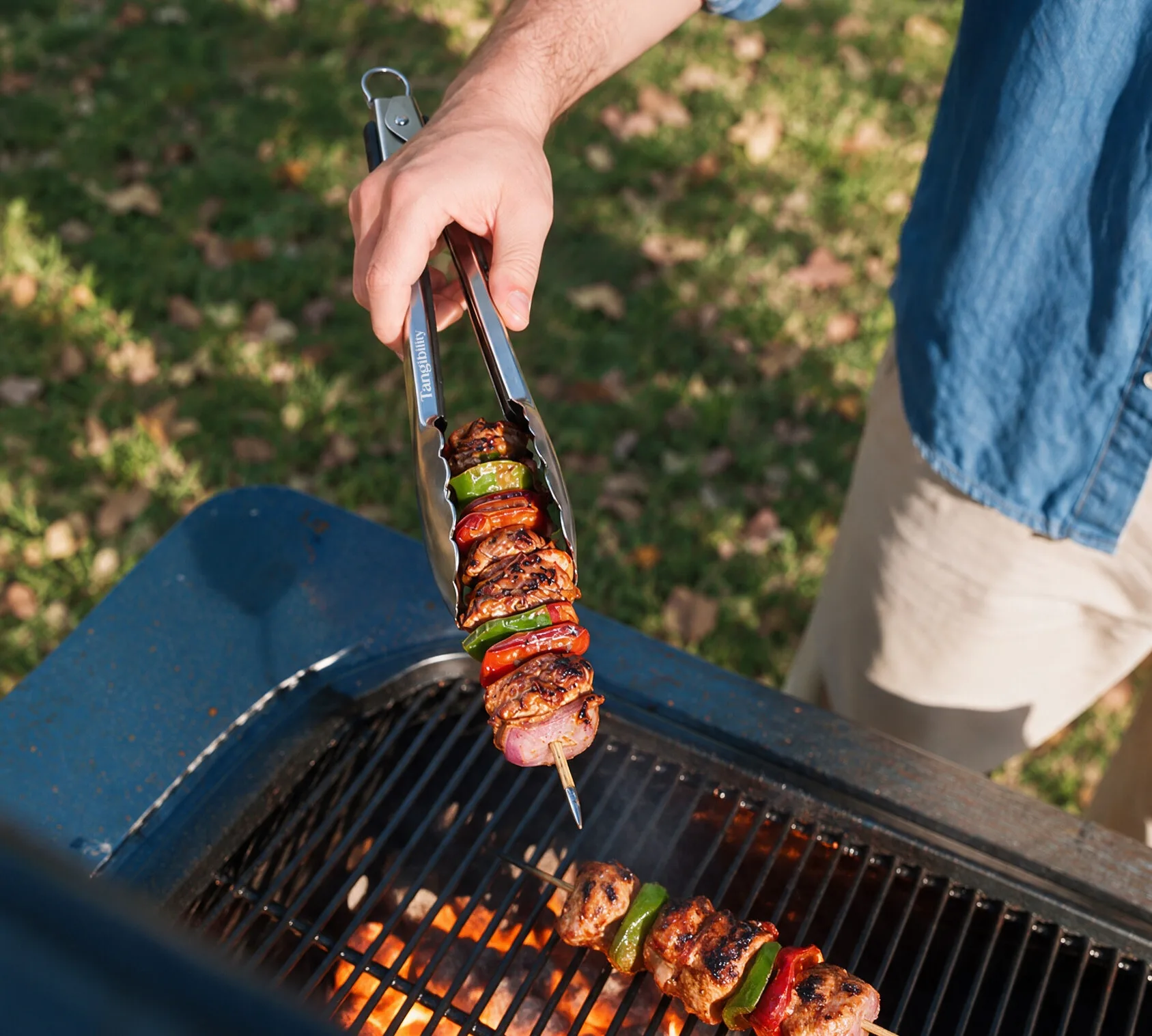 Person using Tangibility tongs to flip a skewer of grilled meat and vegetables over a barbecue.