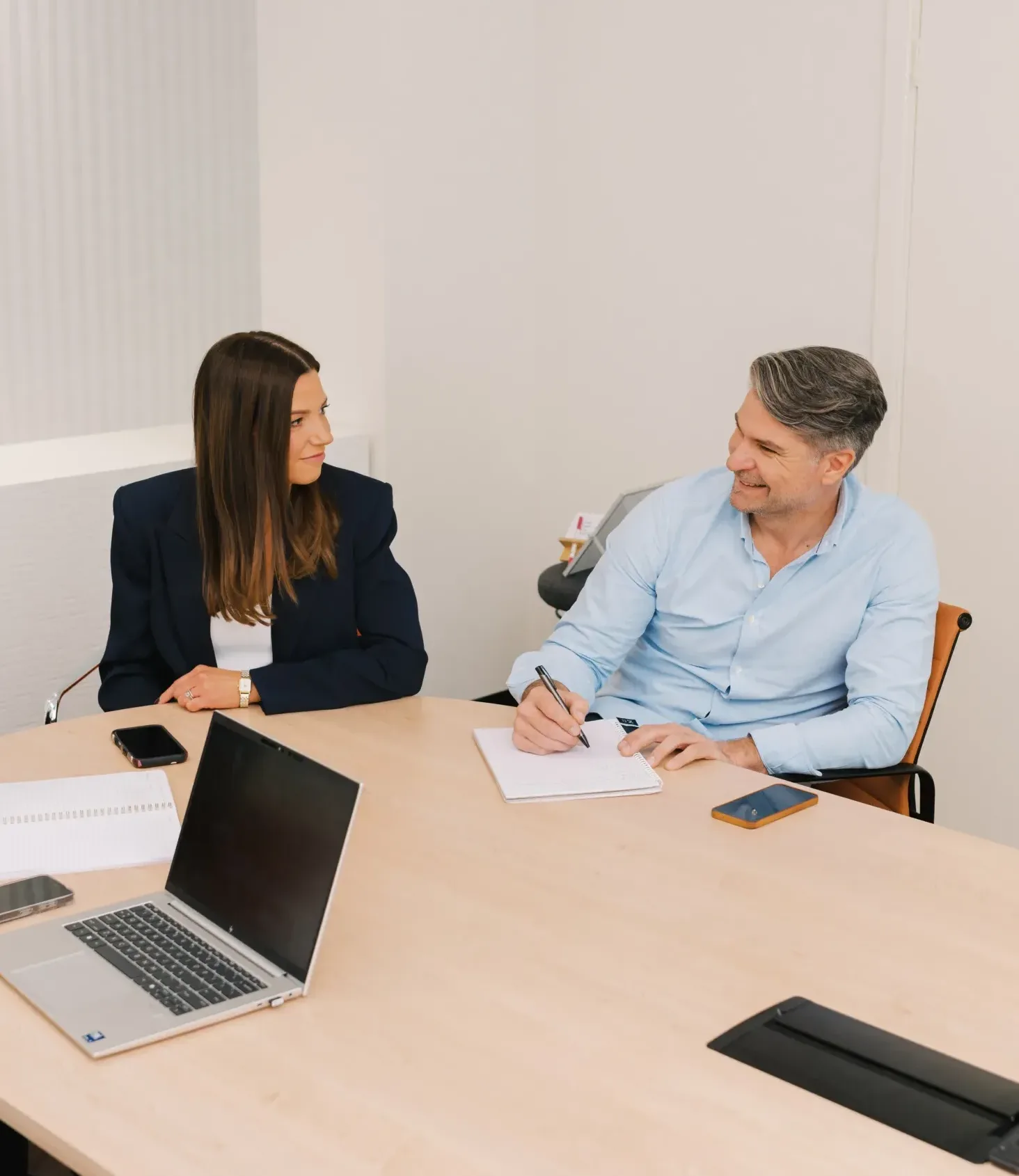 A man and a woman in business attire sit at a conference table discussing work.