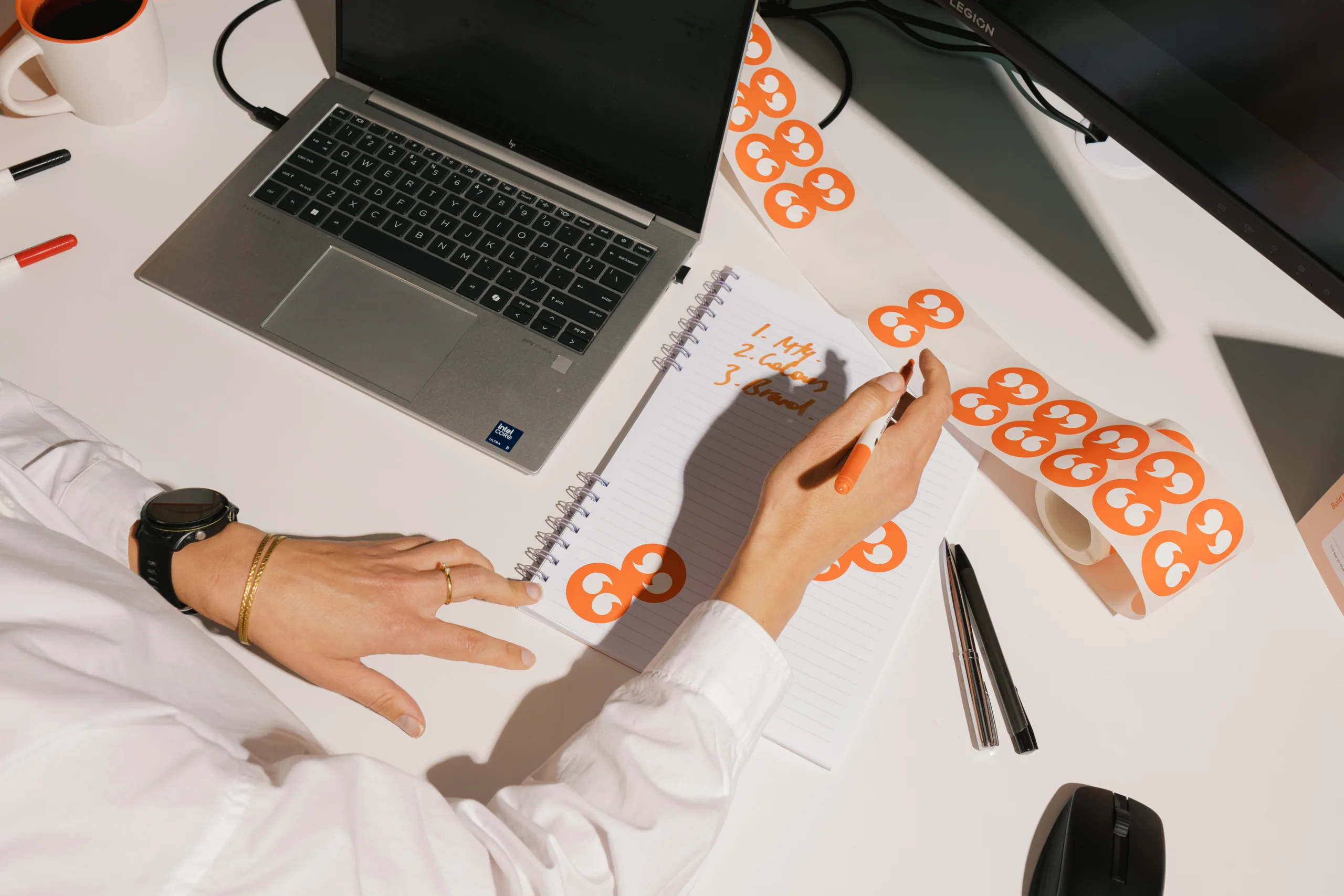 Person writing on a notepad with a laptop and a roll of stickers featuring an orange double quotation mark logo.
