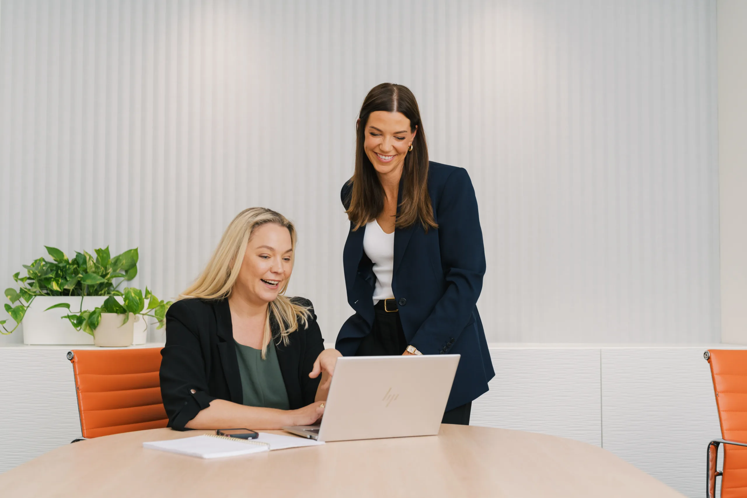 Two women in business attire collaborate over a laptop in a modern office setting.