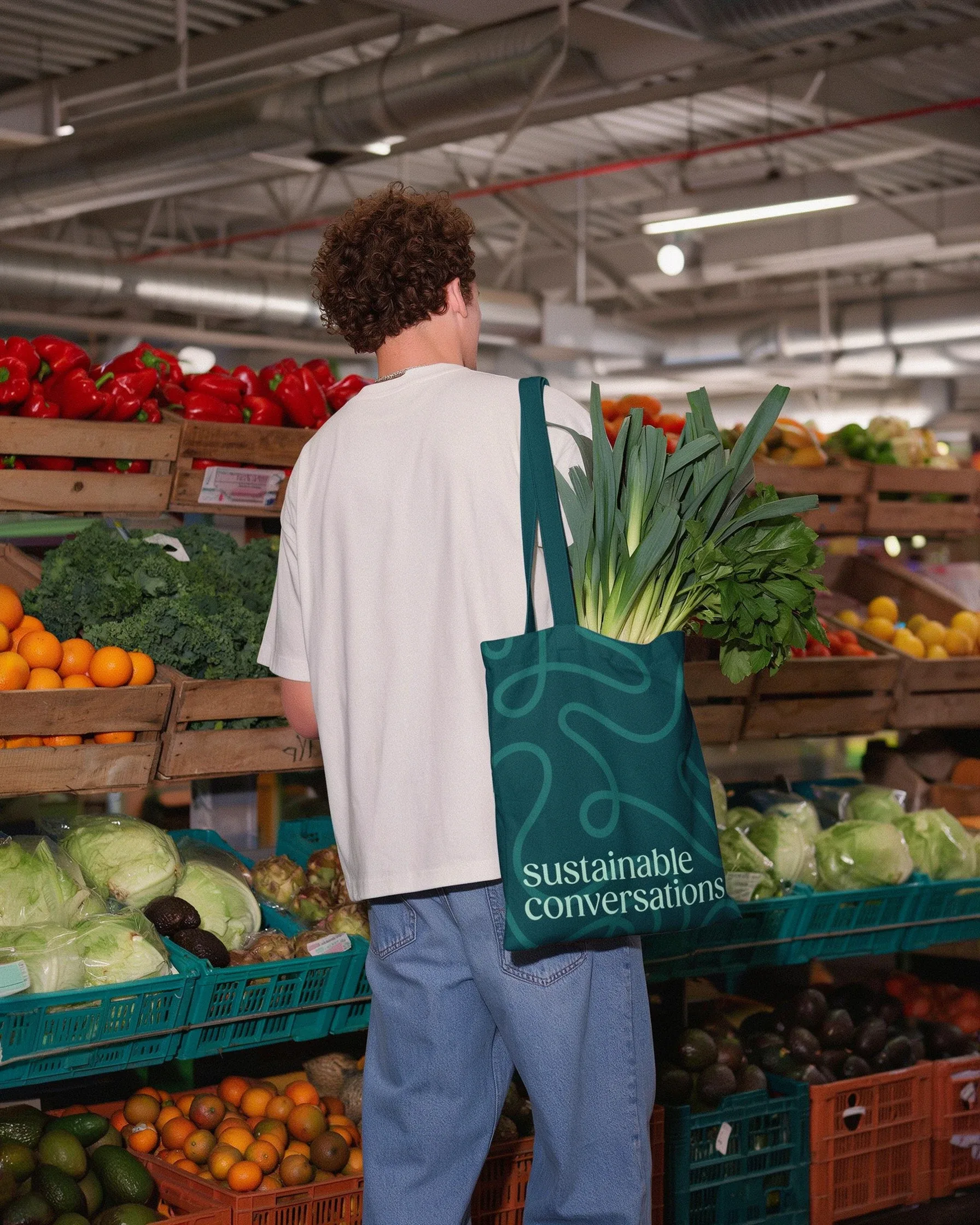 A person carries a teal tote bag with "sustainable conversations" printed on it, filled with leeks and other greens, at a
