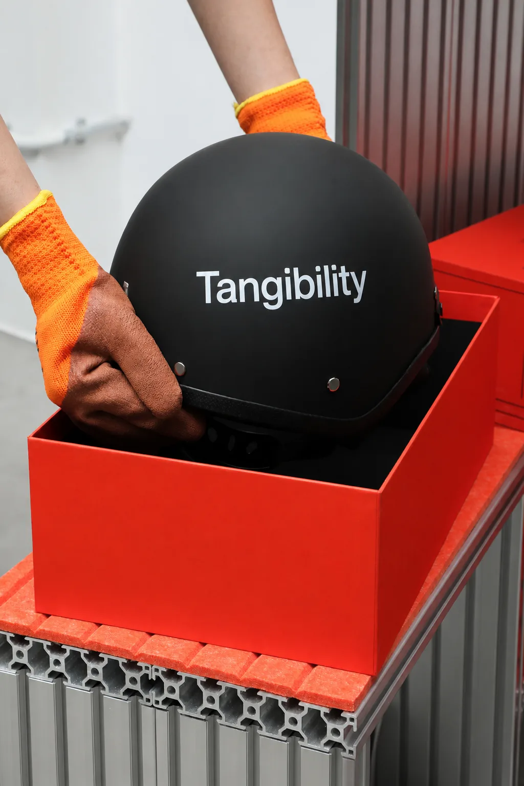 A black helmet with "Tangibility" printed in white is placed into a bright orange box by hands wearing orange gloves.