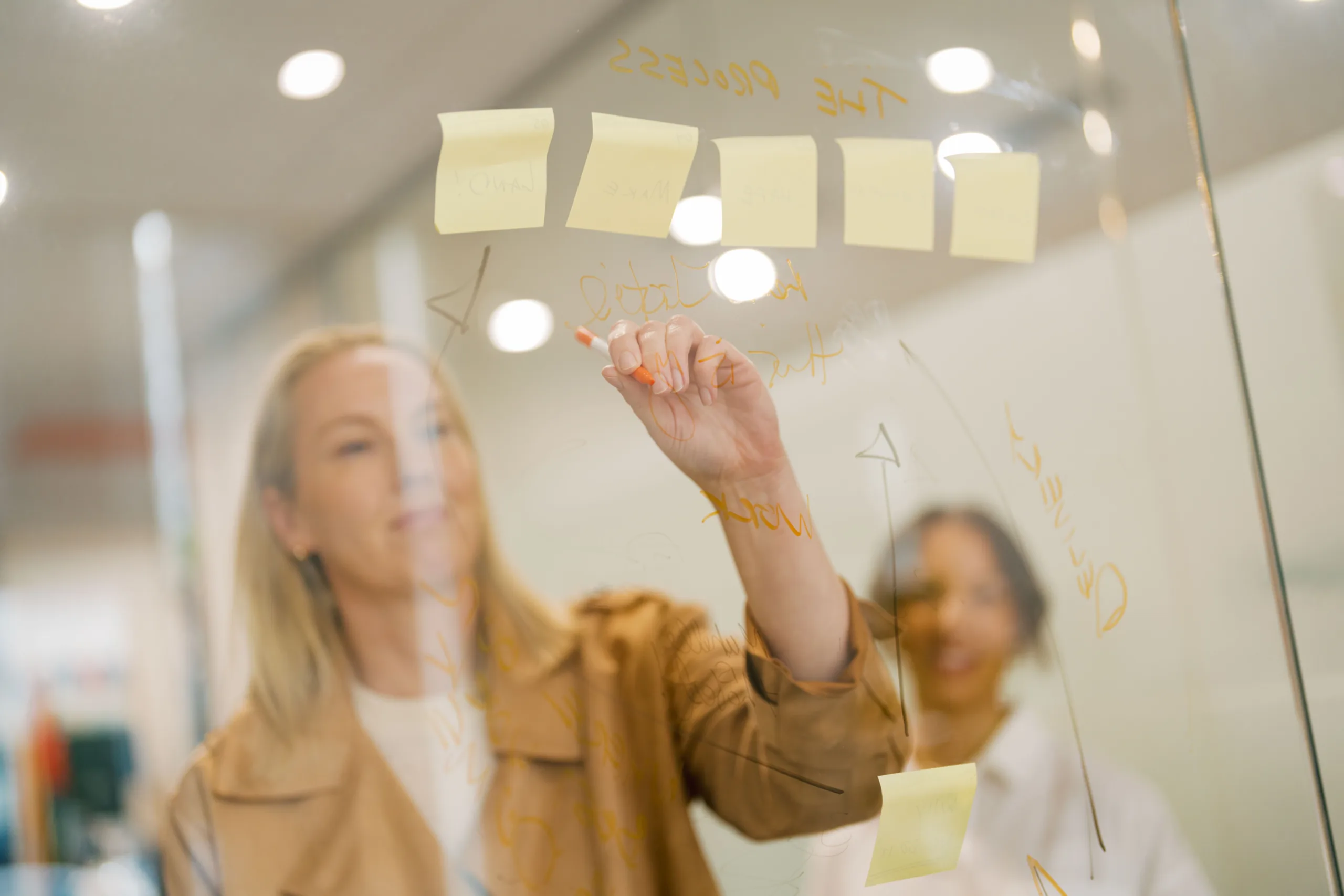 Two women collaborate on a glass board, writing and placing sticky notes.