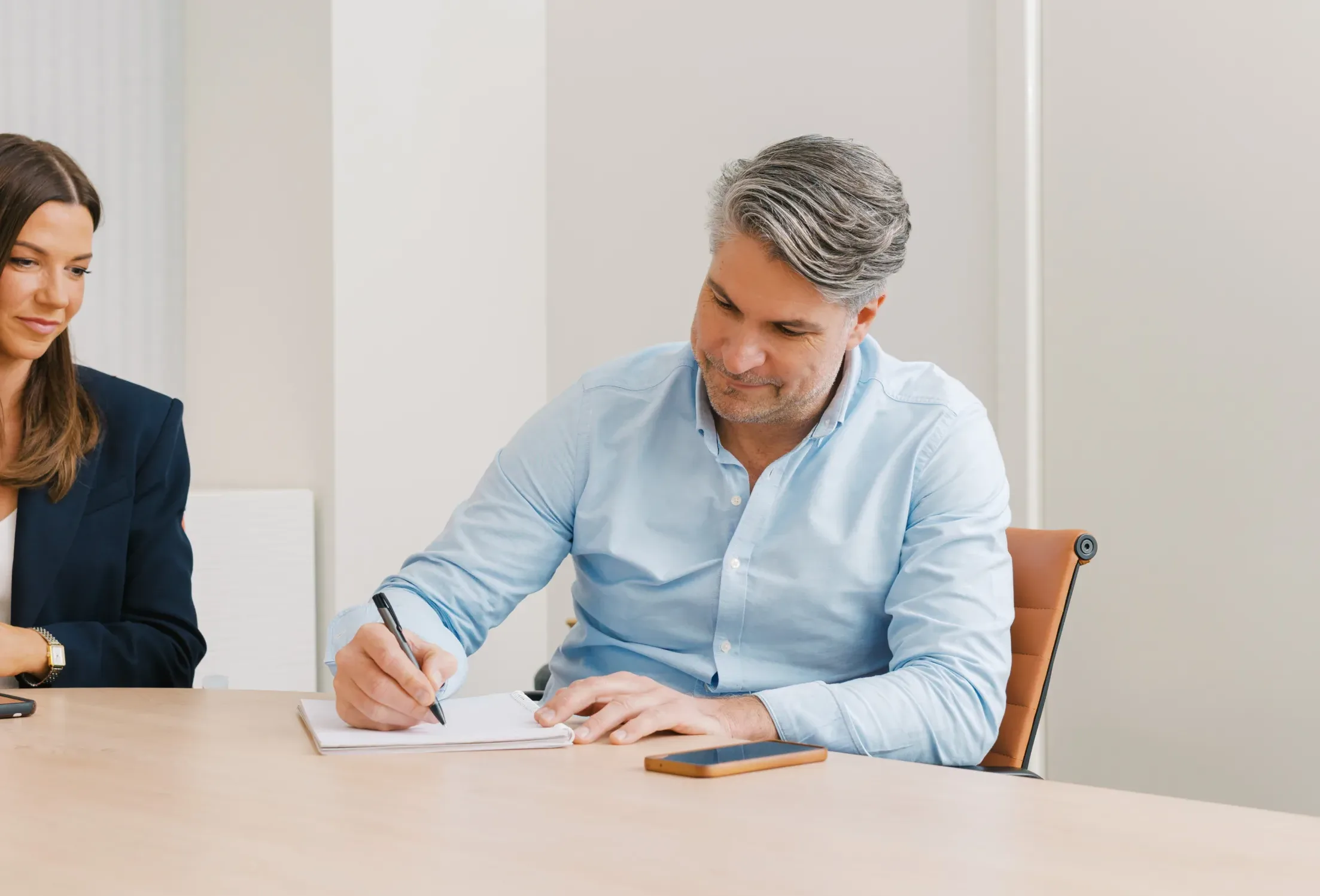 A man in a light blue shirt writes in a notebook at a table while a woman in a dark blazer looks on.