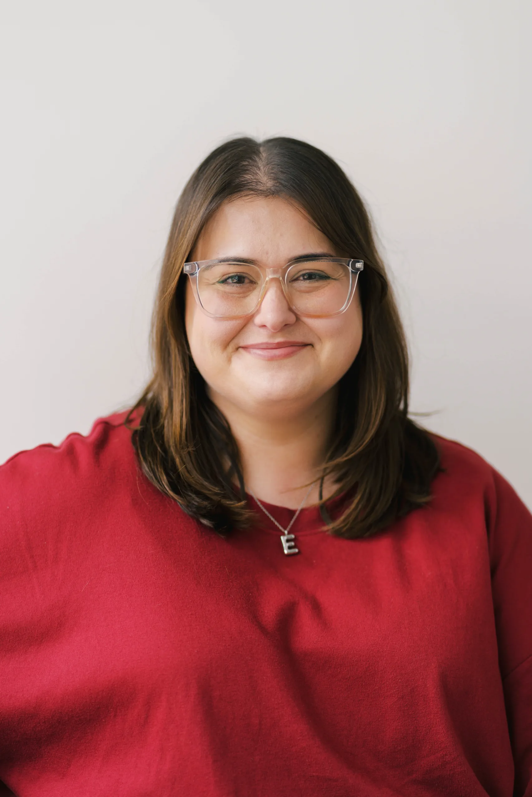 Gemma McCrory wears glasses and a red shirt with a silver necklace featuring the letter E.