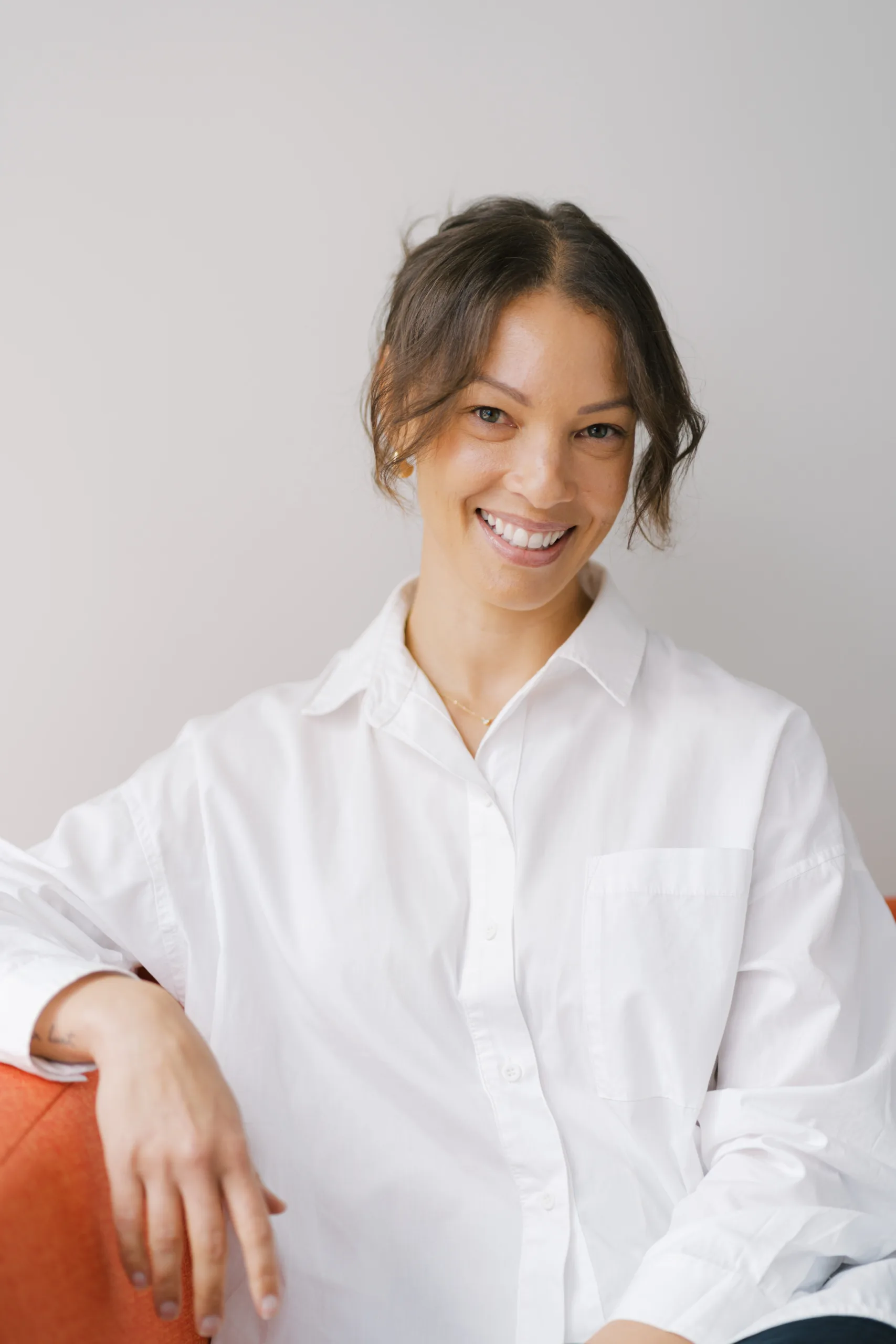 Jacqui Zanetti smiles warmly while wearing a white button-down shirt and leaning on an orange couch.