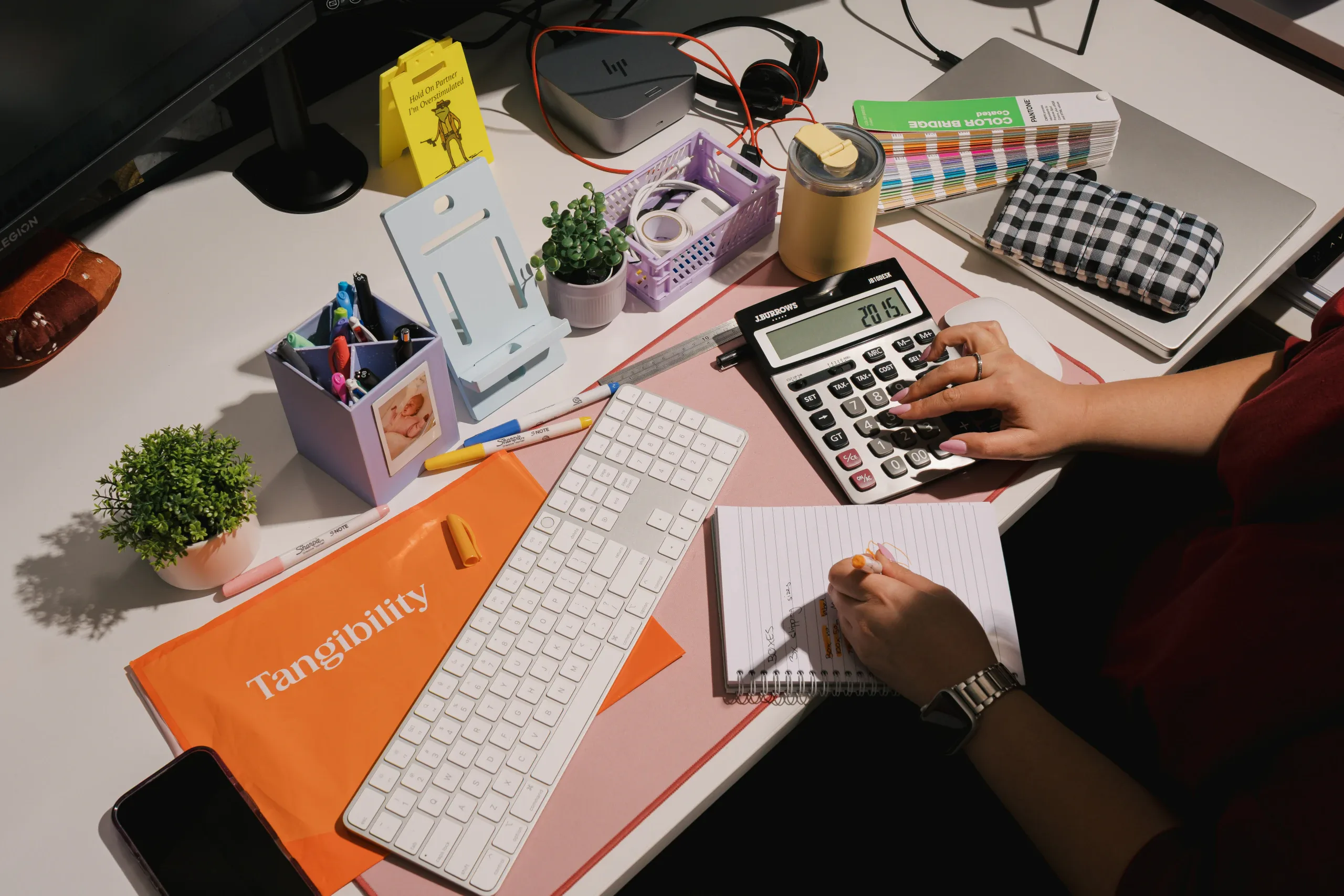 A person uses a calculator and notebook at a desk with Tangibility branding and office supplies.
