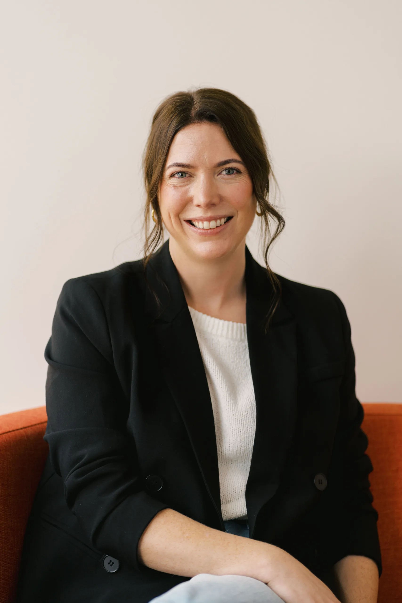 Gemma McCrory smiles while wearing a black blazer and white sweater, seated on an orange couch.