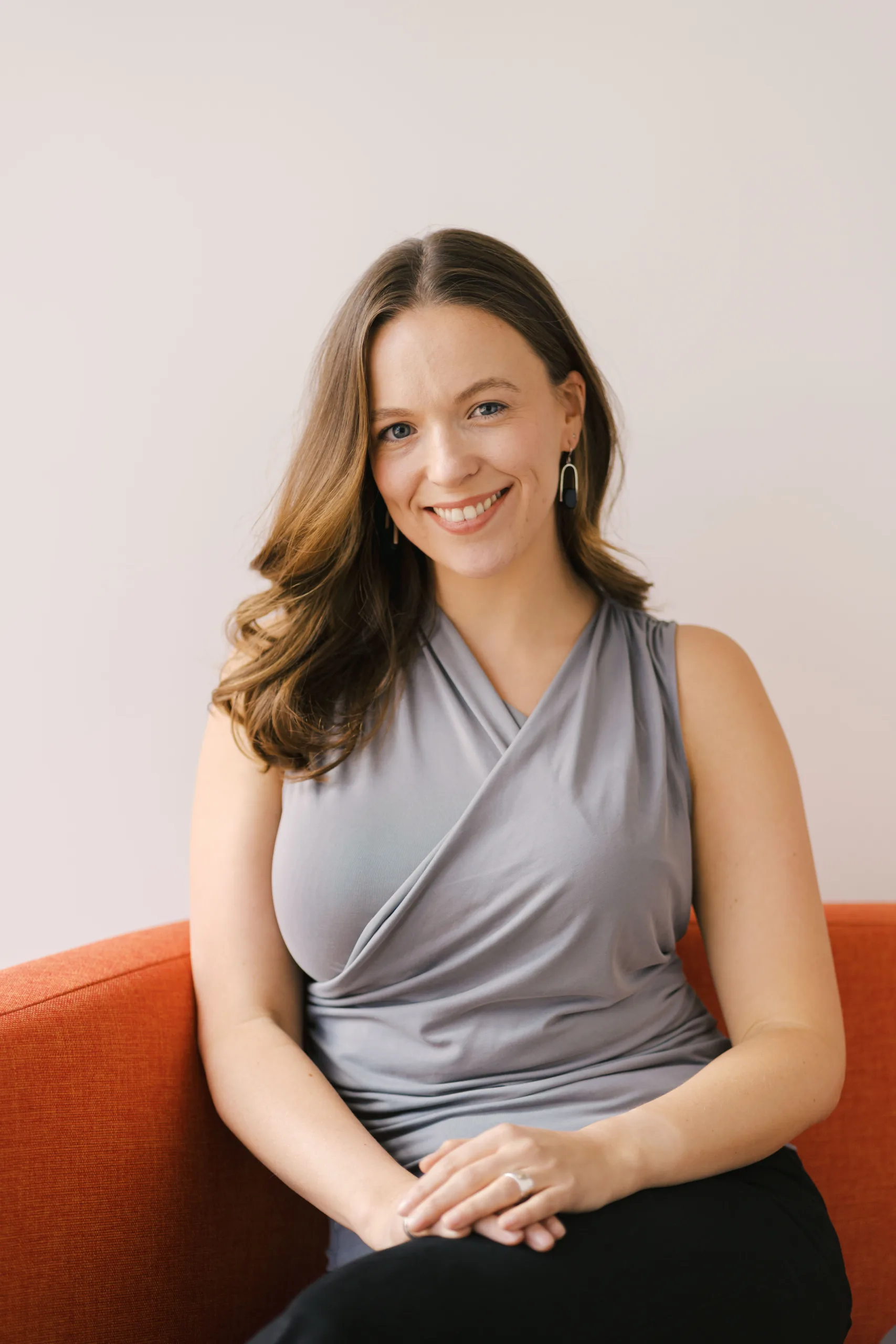 A woman with brown wavy hair smiles while sitting on an orange couch.