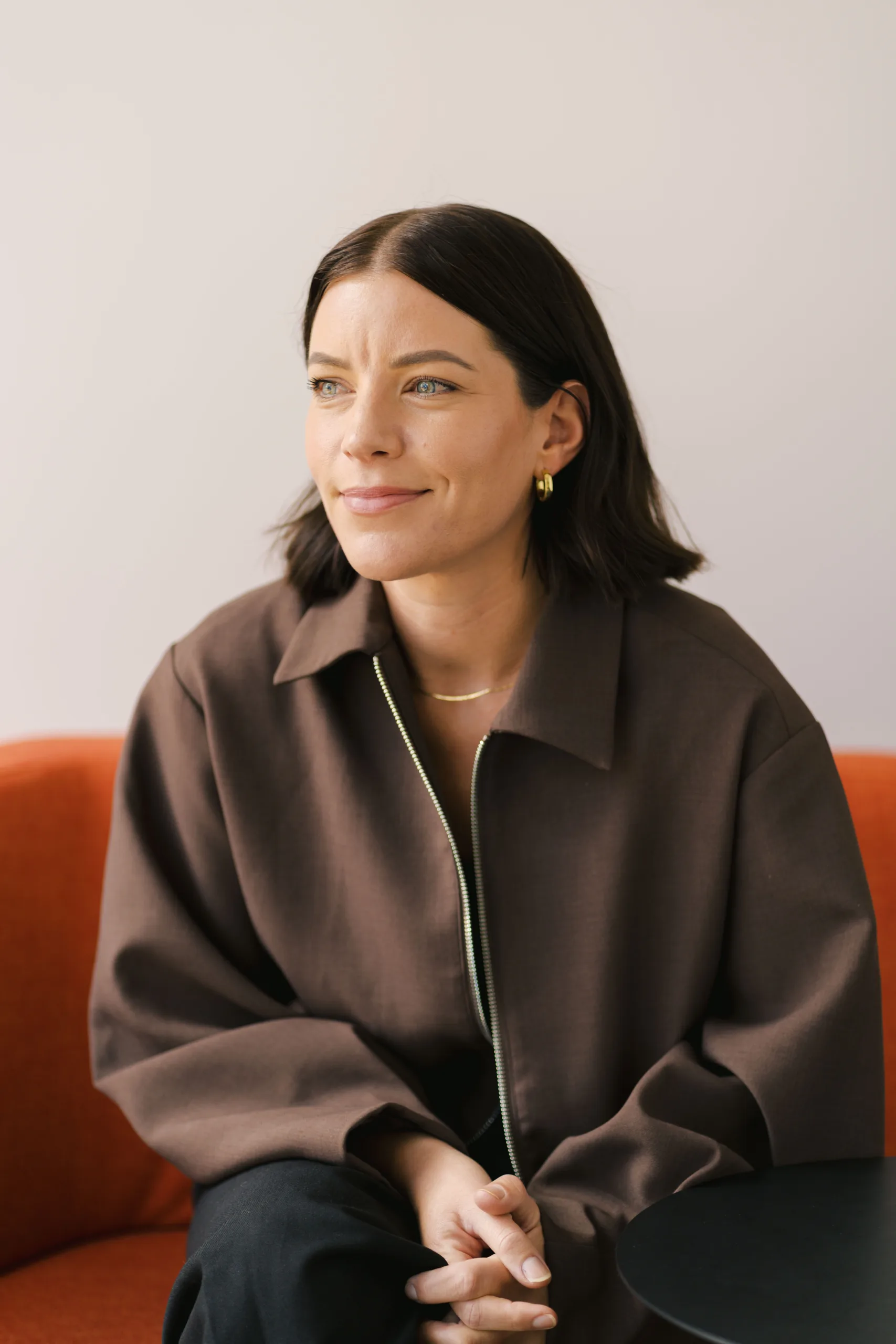 Amy Grubb smiles and looks to the side while wearing a brown jacket and gold earrings.
