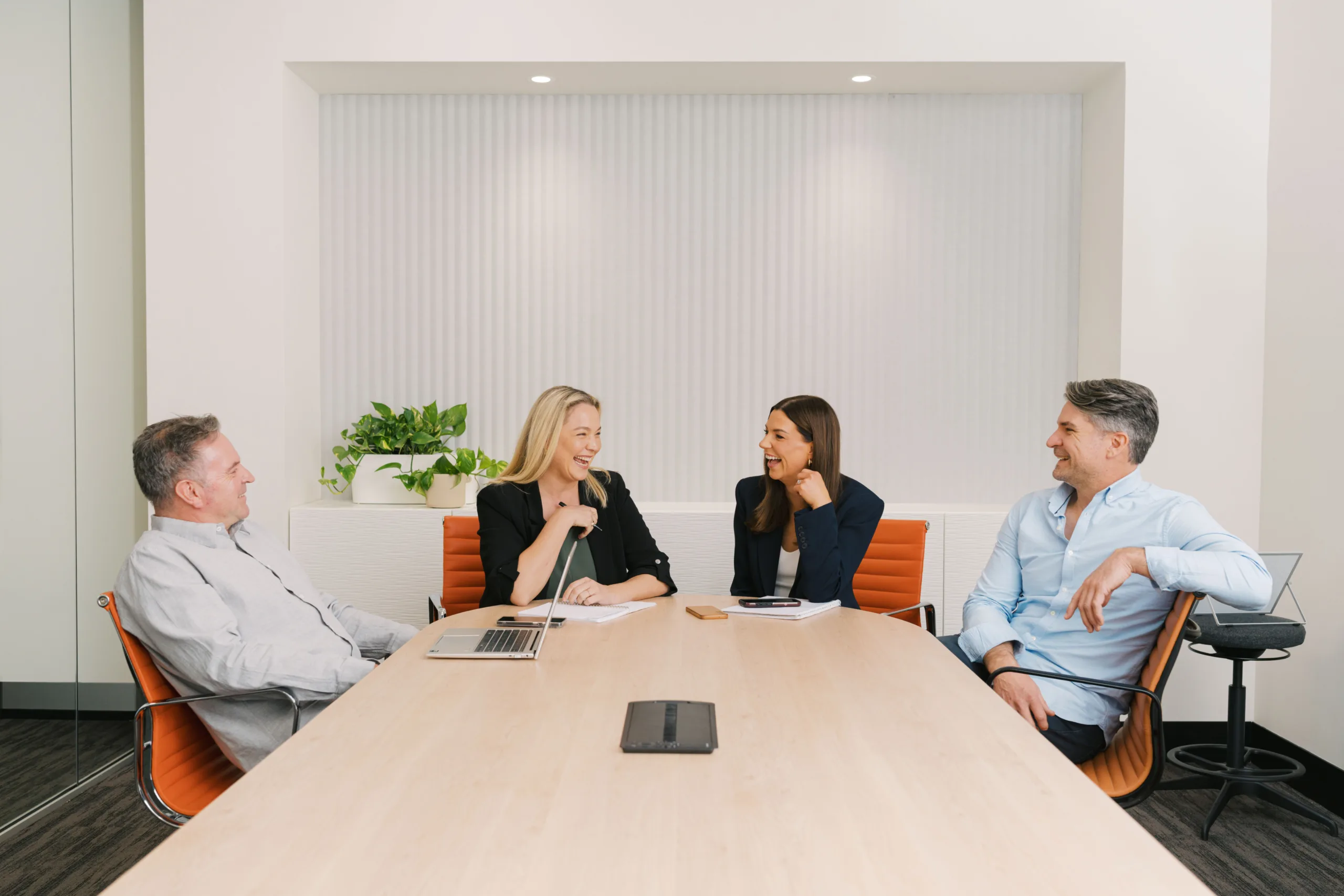 Four professionals in a modern office setting, seated around a conference table and engaged in conversation.