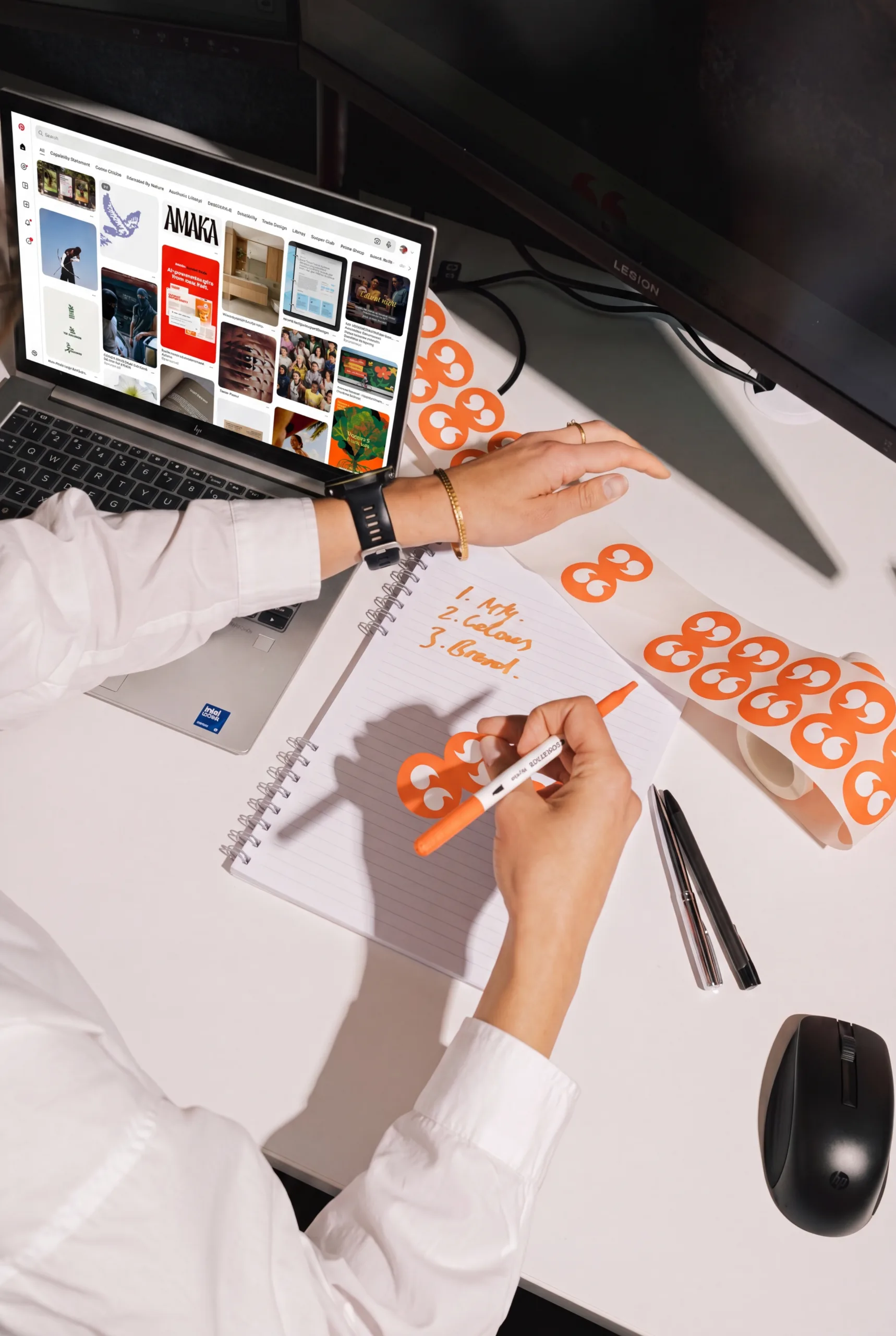Person writing on a notepad with a laptop and roll of branded stickers nearby.