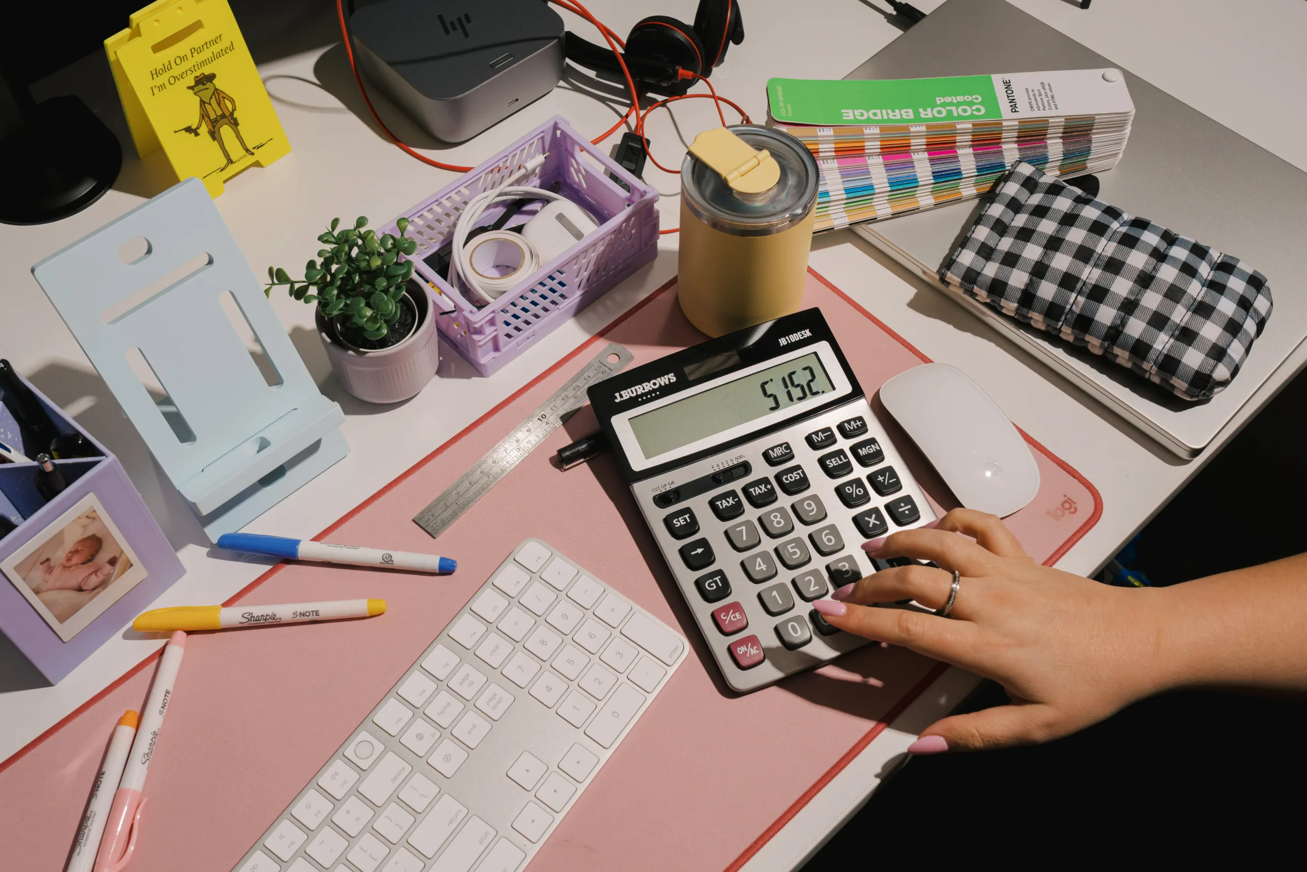 A hand uses a J. Burrows calculator displaying 5152 on a desk with a keyboard, mouse, and color swatches.