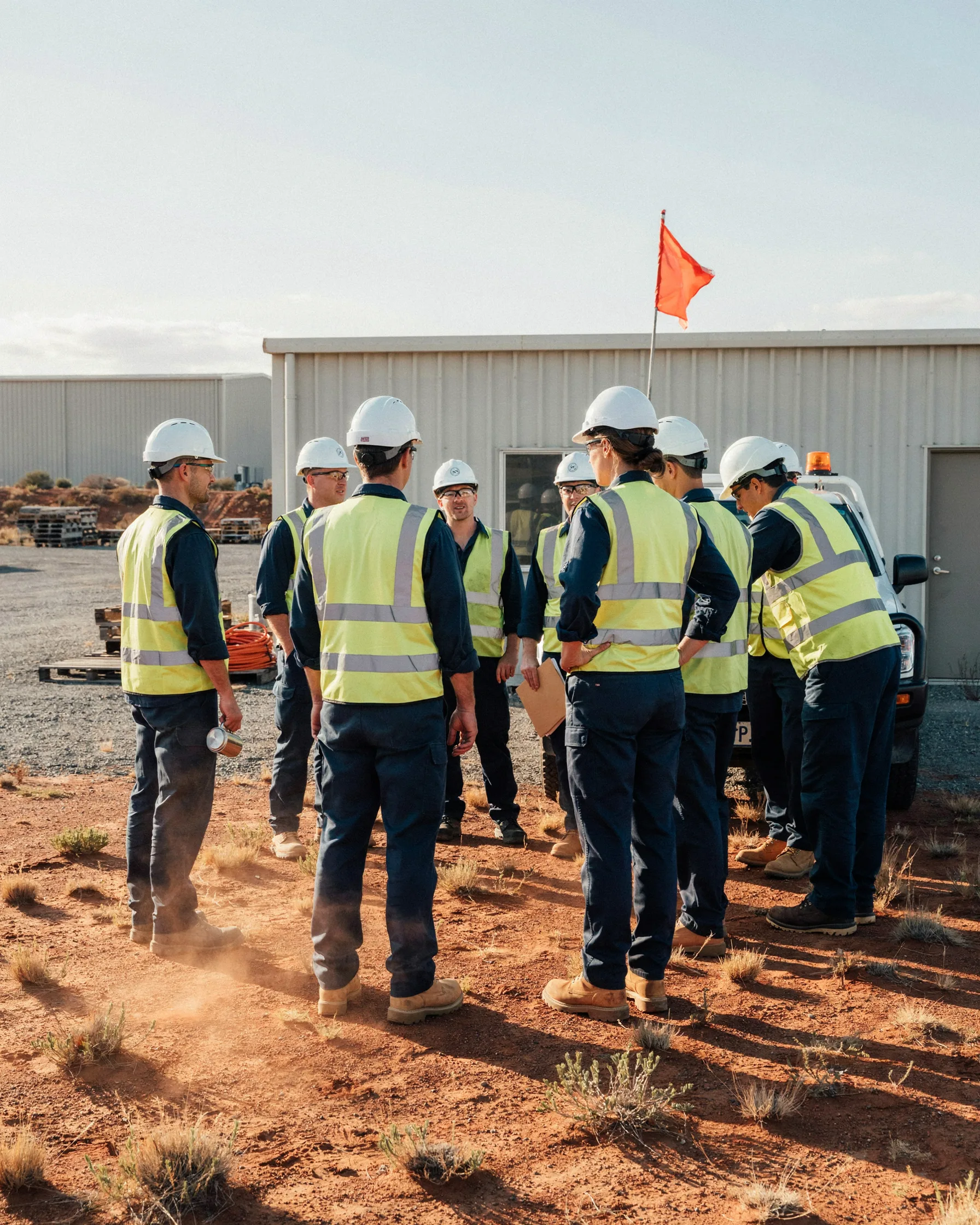 A group of workers in hard hats and high-visibility vests stand in a huddle outdoors near a building.