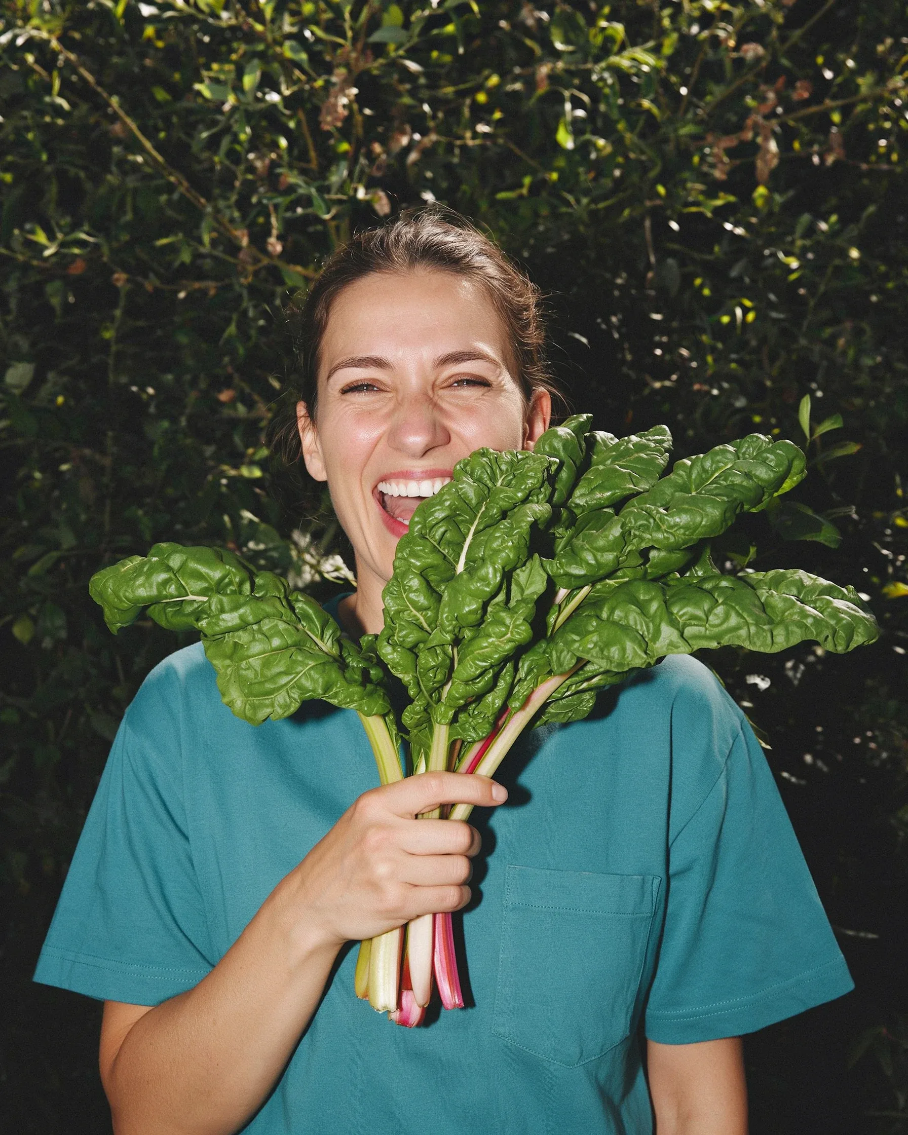 A laughing woman holds a bunch of rainbow chard in front of her face against a dark green leafy background.