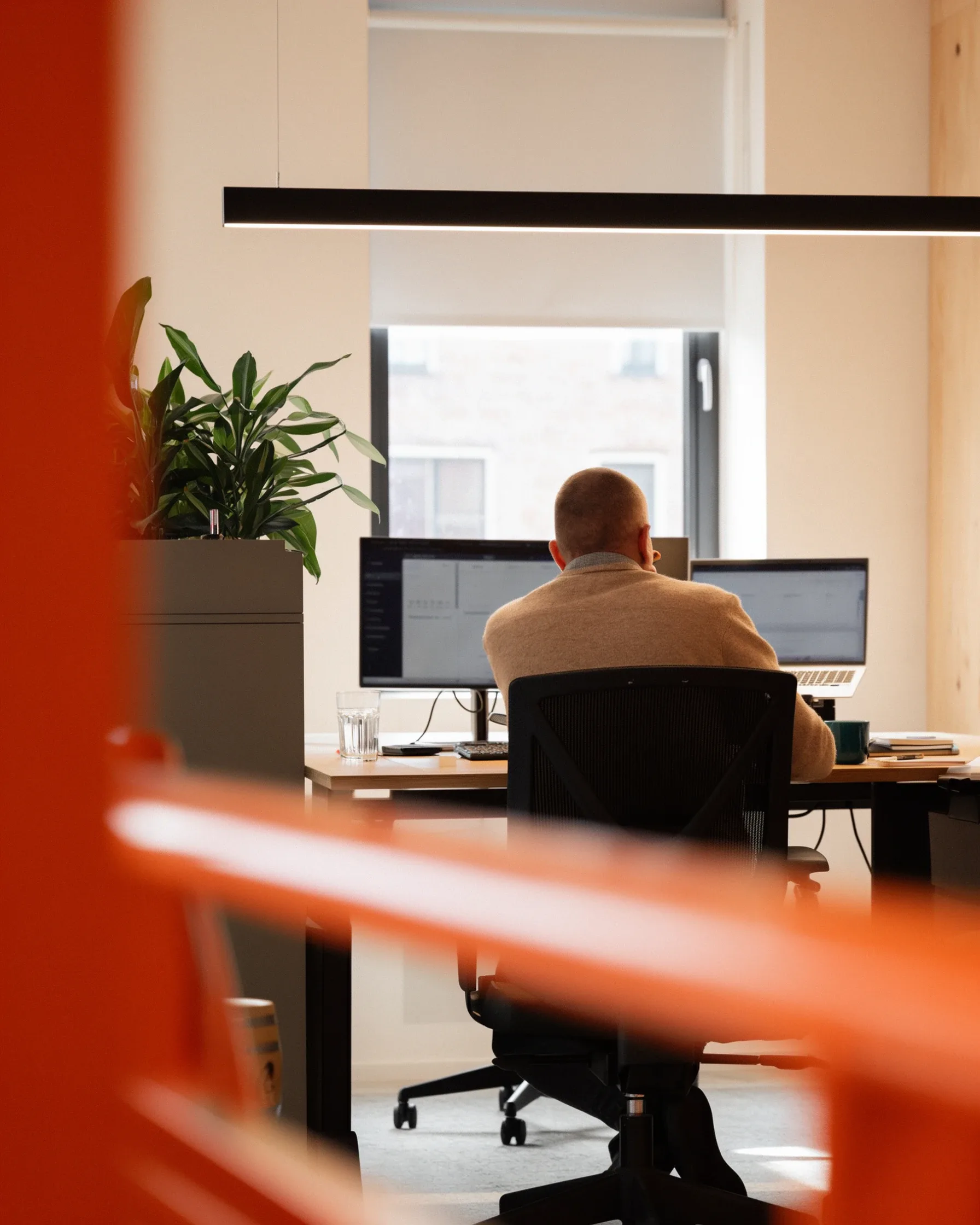 Man in a tan sweater works at a desk with dual monitors in a modern office.