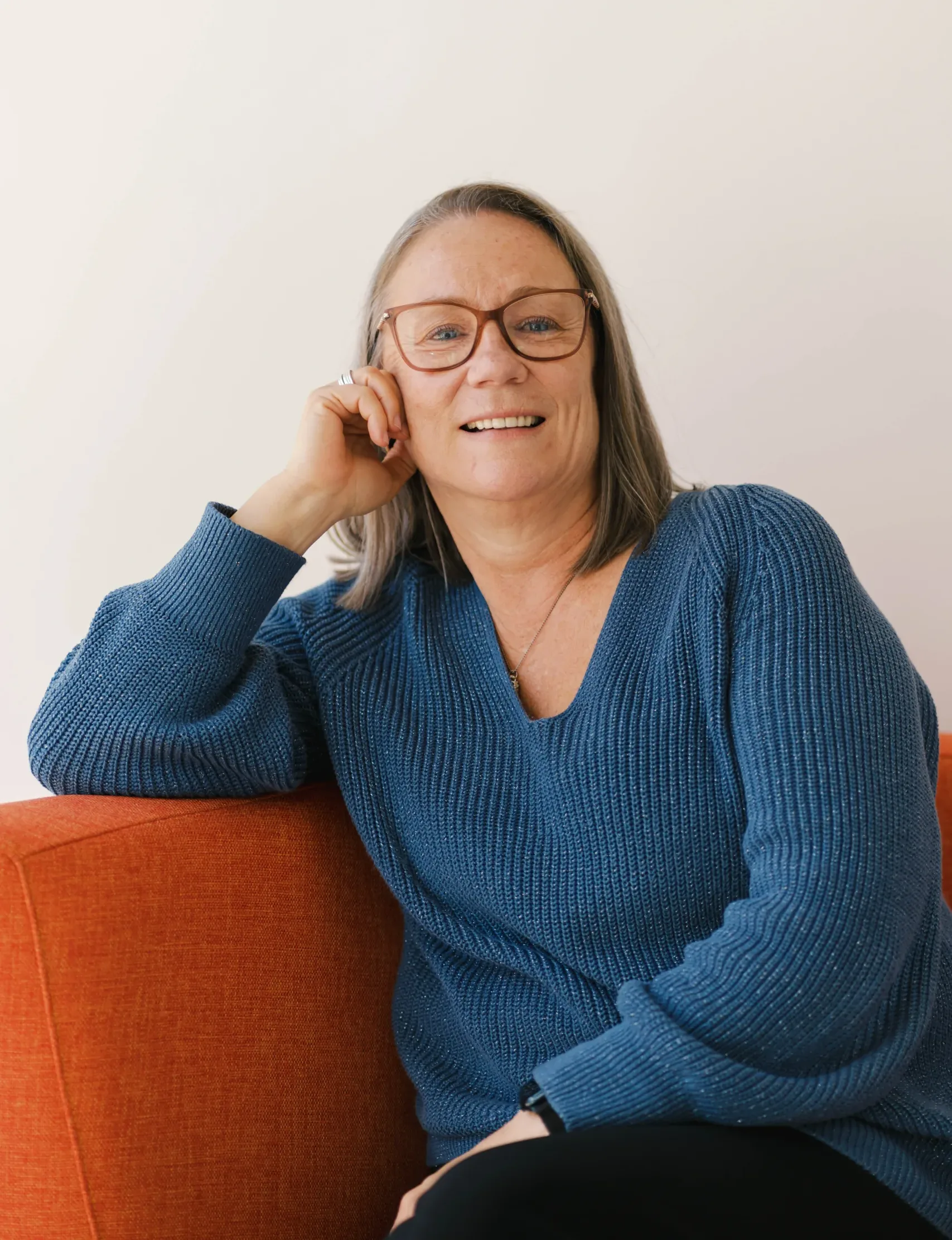Lisa Ammon smiles while wearing glasses and a blue sweater, leaning on an orange couch.