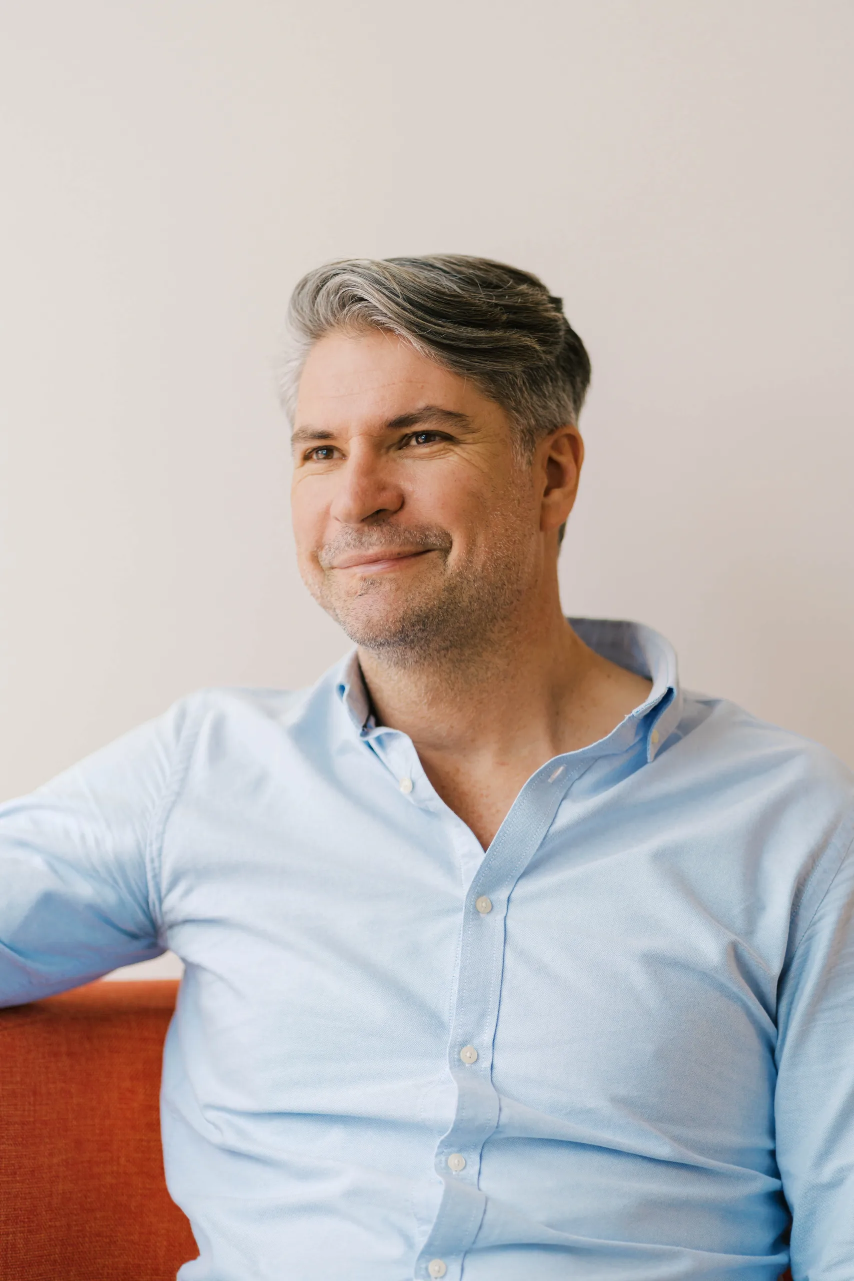 Jared Schipper smiles while wearing a light blue button-down shirt and sitting on an orange couch.
