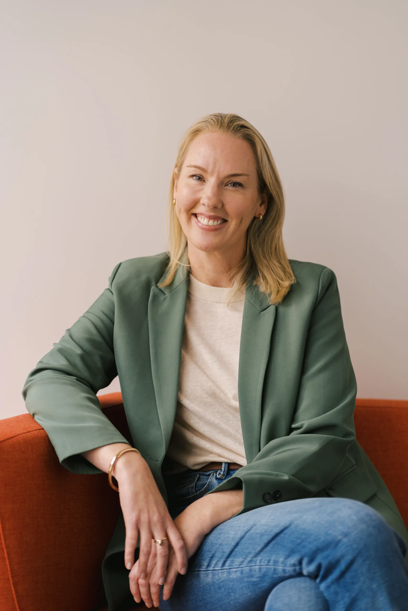 Michelle Tickle smiles while wearing a green blazer and jeans, seated on an orange couch.