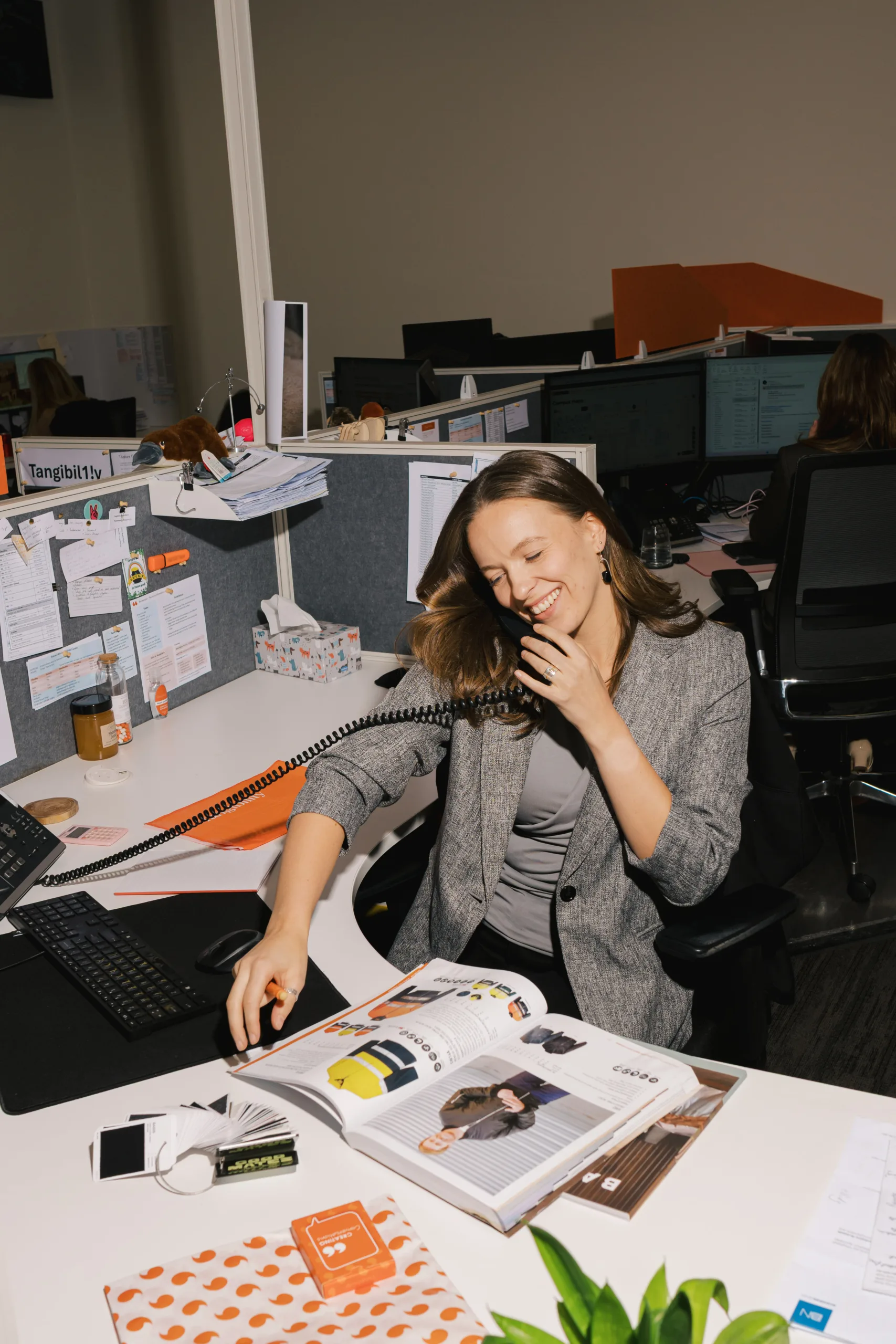 Woman in a grey blazer smiles while talking on a corded phone at a desk with an open magazine.