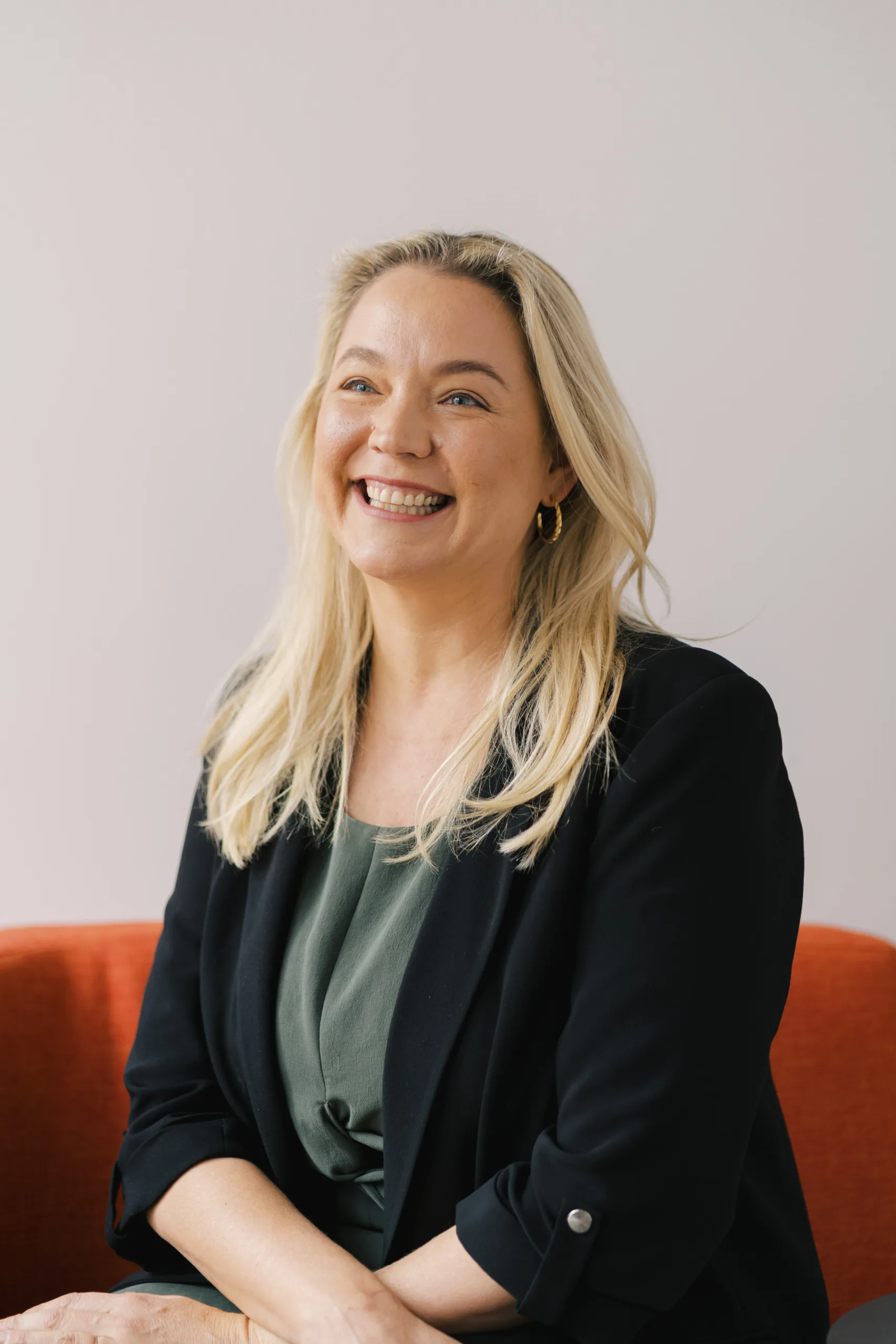 Lisa Ammon smiles while wearing a black blazer and green top, seated on an orange couch.