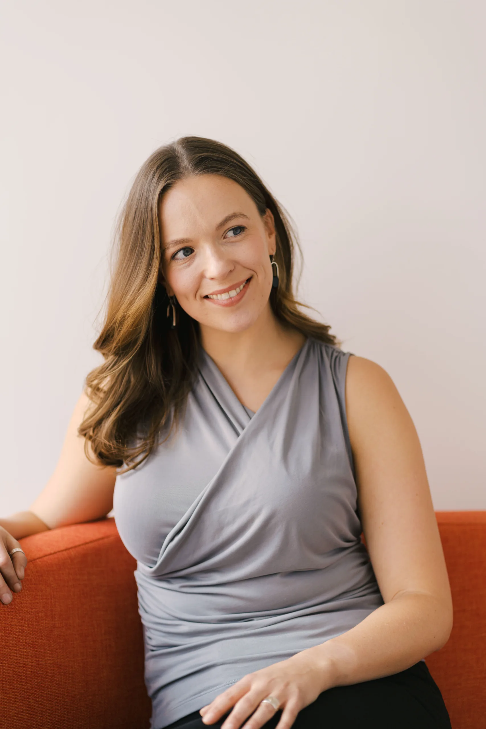 A woman with long brown hair smiles and looks to the side while sitting on an orange couch.