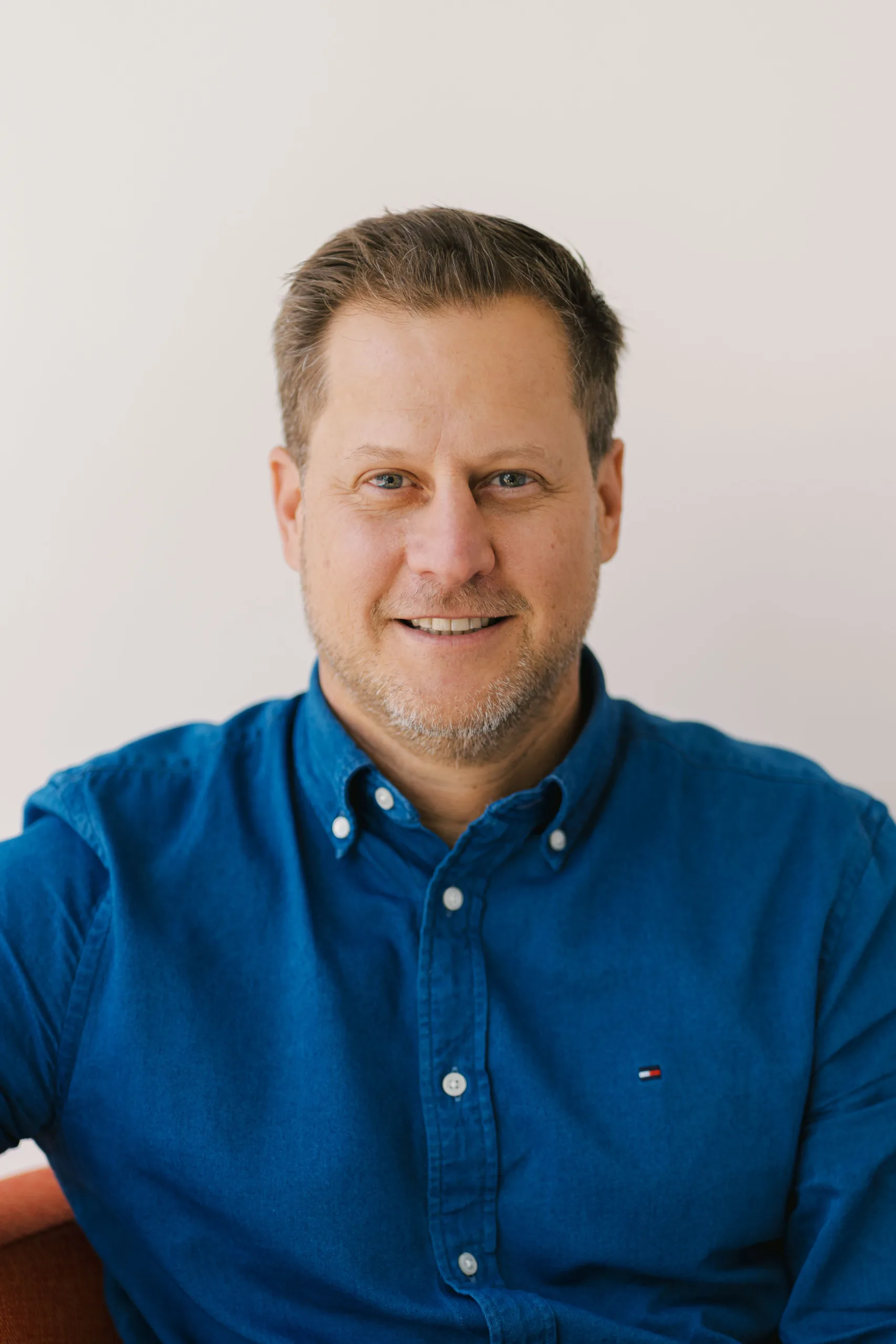 John Tasker smiles wearing a blue Tommy Hilfiger shirt against a white background.